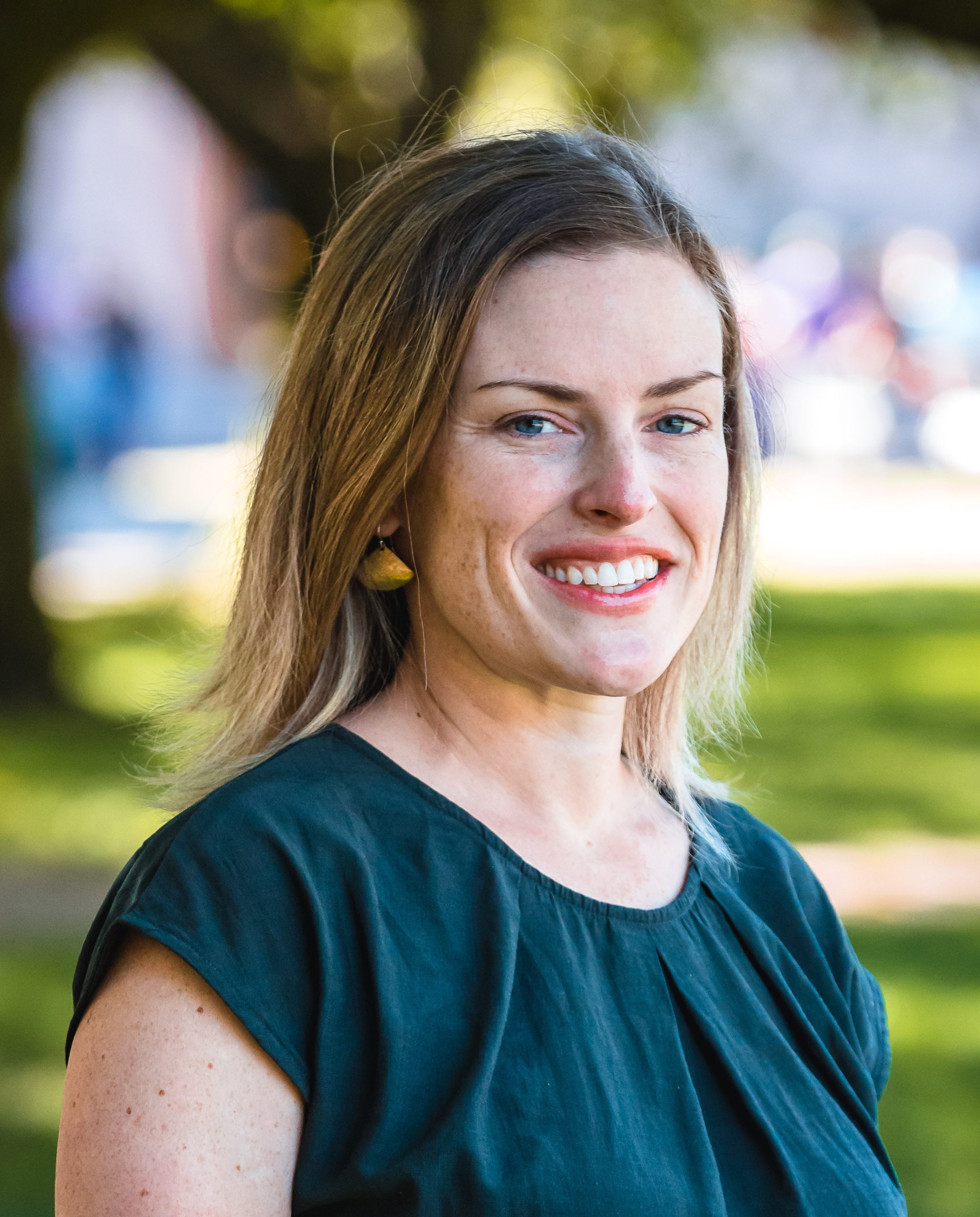 White woman with straight hair smiling wearing gold earrings and a teal shirt