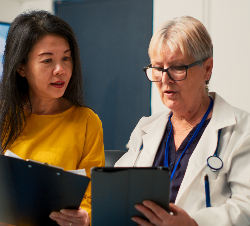 An Asian patient has a clipboard in her hand and is looking at a tablet an older white woman doctor is holding. The doctor is explaining something to the patient. 