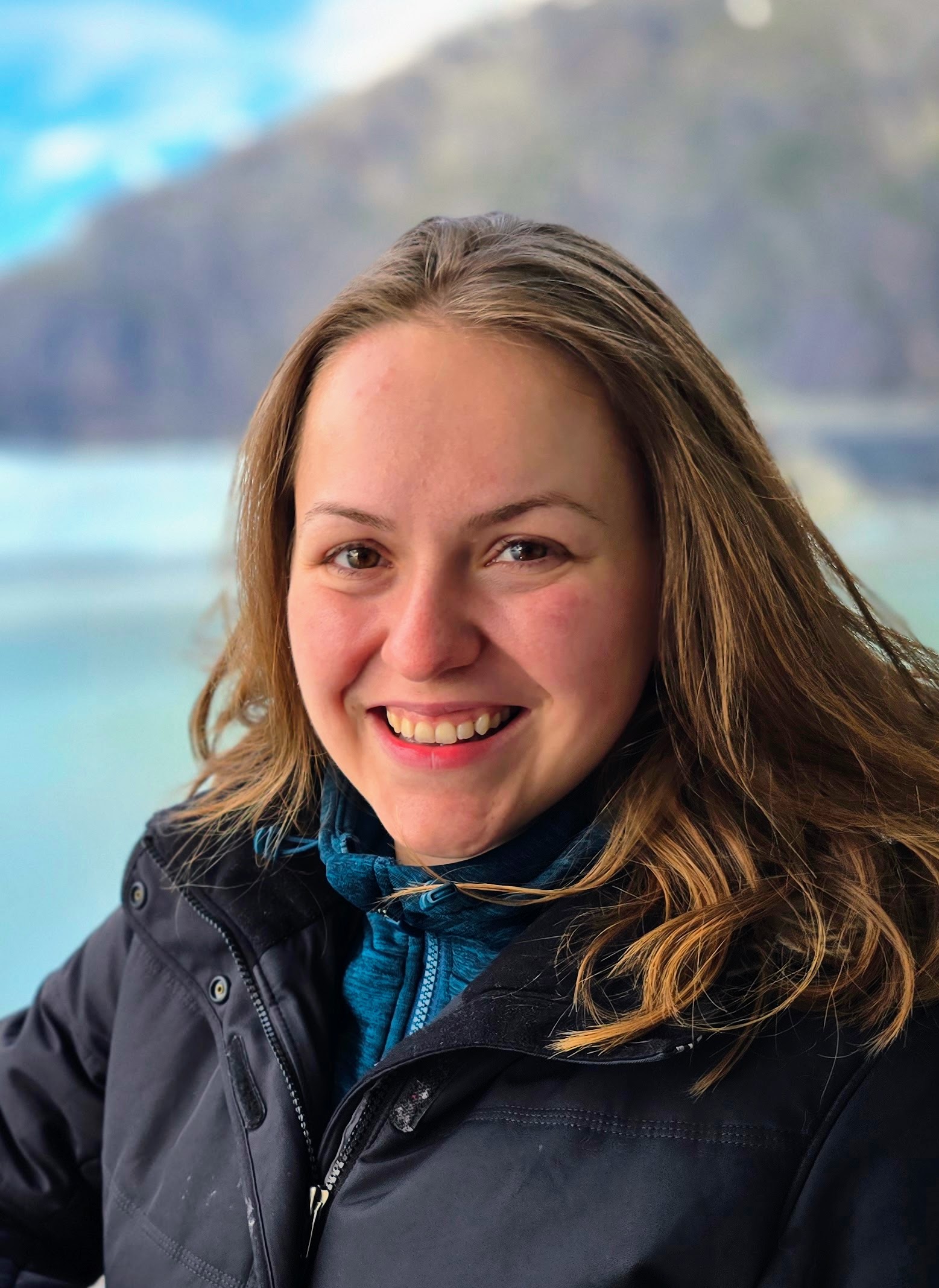A headshot of a young white woman with shoulder length light-brown hair, a black coat, against a blue and brown background.
