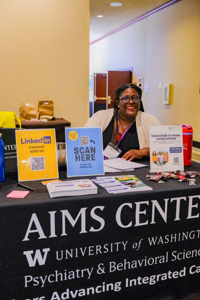 Monica Harrison smiling from behind a table with an AIMS Center table cloth and swag at a recent conference. 