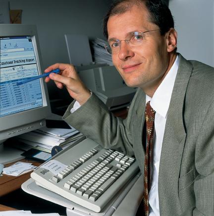 A young Dr. Unützer, a light skinned man wearing glasses, points with a pen to a computer screen with the first AIMS Caseload Tracker on the screen.